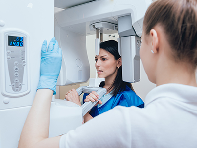 A woman in a blue lab coat is standing next to a large, modern 3D scanner with a digital display showing the word scan and a timer reading 0 42, while another person in a white lab coat is observing the scan.